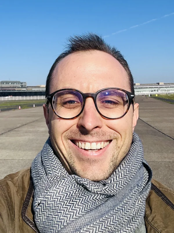 Tamas smiling in glasses and a scarf on a sunny airfield