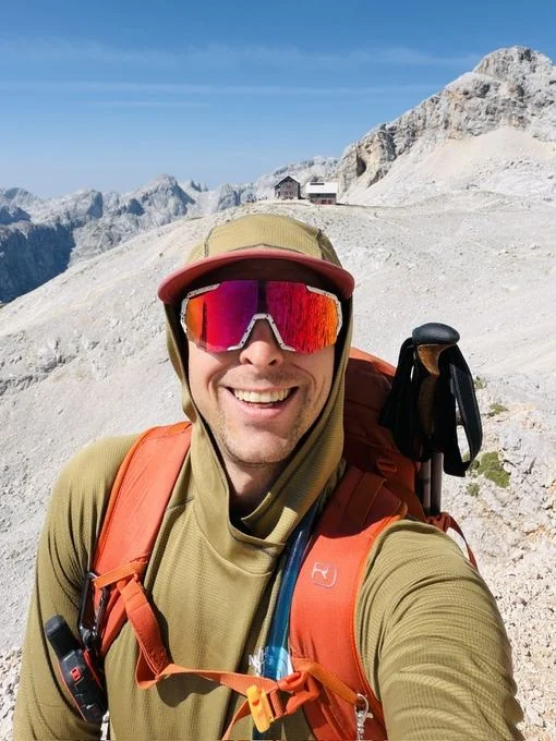 Tamas, smiling in sunglasses and a cap on a rocky mountain ridge, with a mountain hut behind him