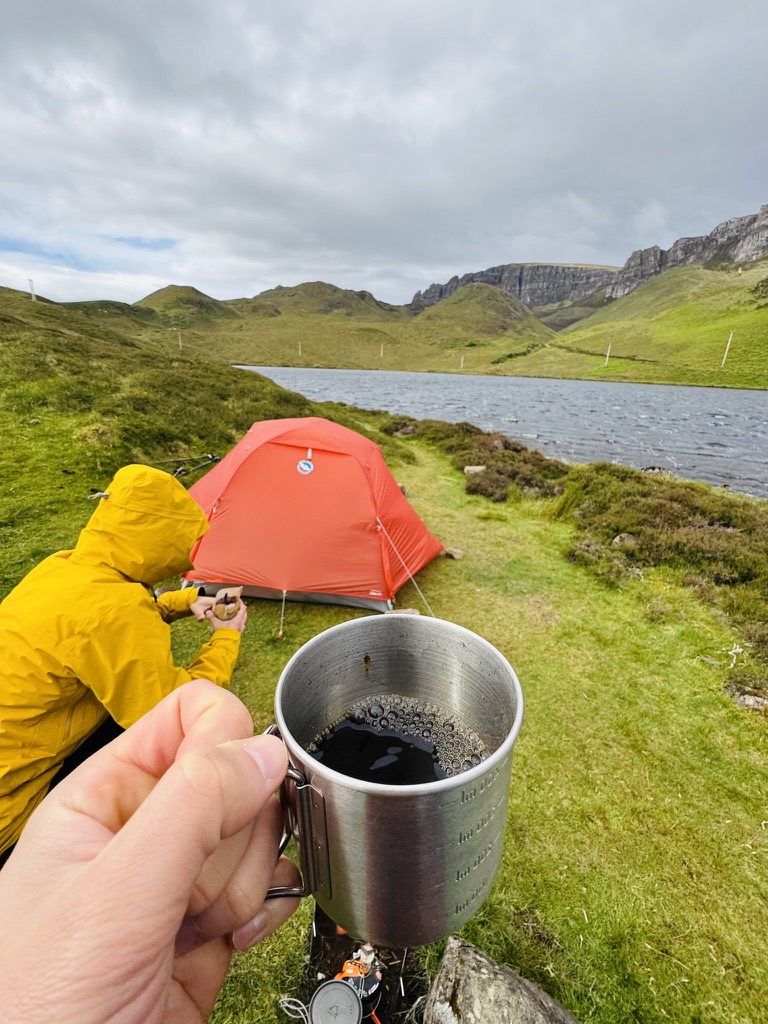 Morning coffee beside an orange tent pitched above a small mountain lake