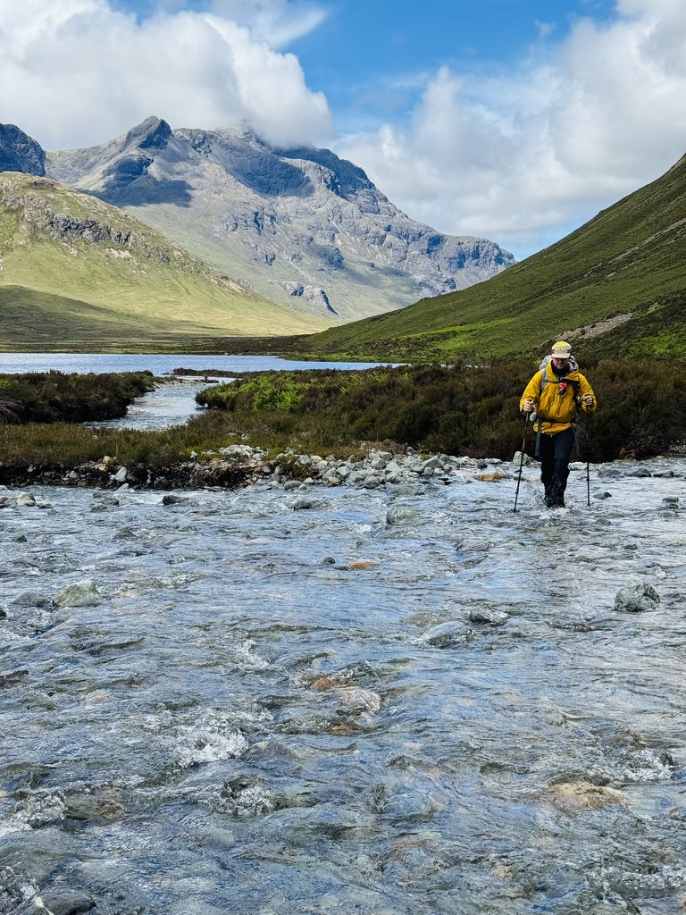 Hiker in a yellow jacket wading a rocky river crossing beneath rugged peaks