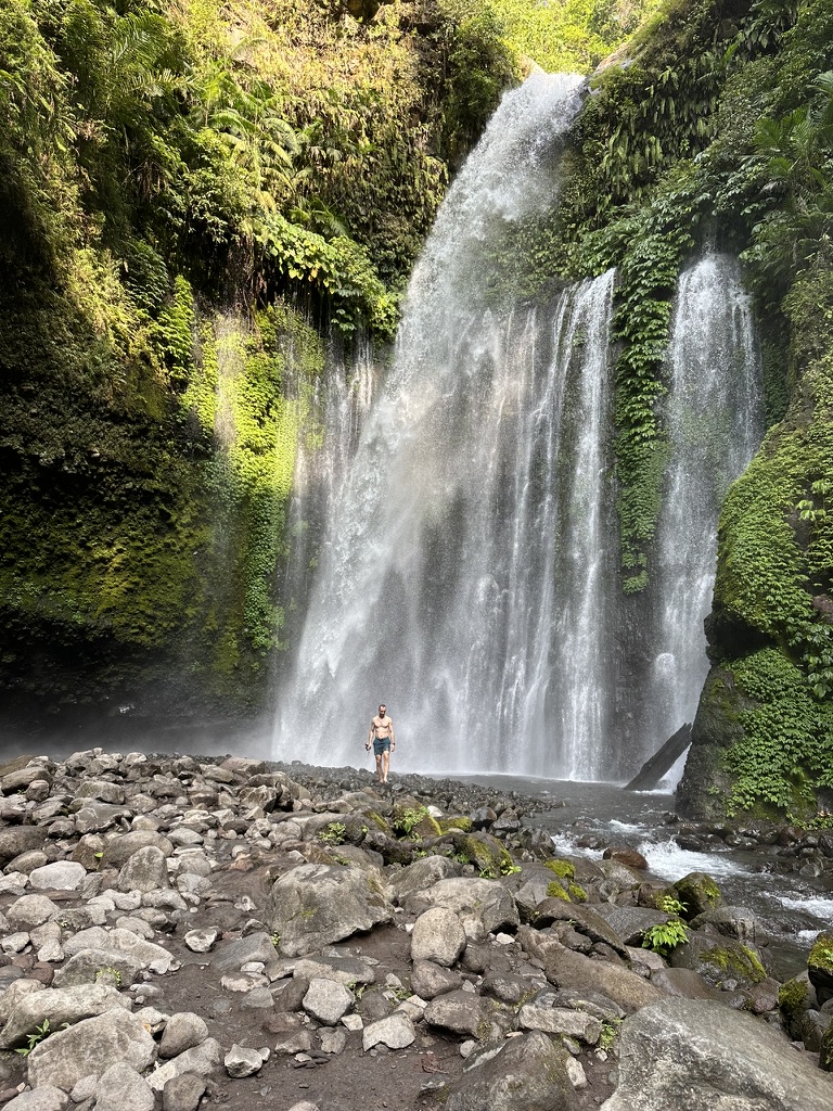 Traveler standing beneath a broad waterfall surrounded by tropical greenery